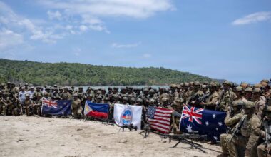 A large number of military personnel dressed in various camouflaged uniforms pose for a photo with flags from various countries.