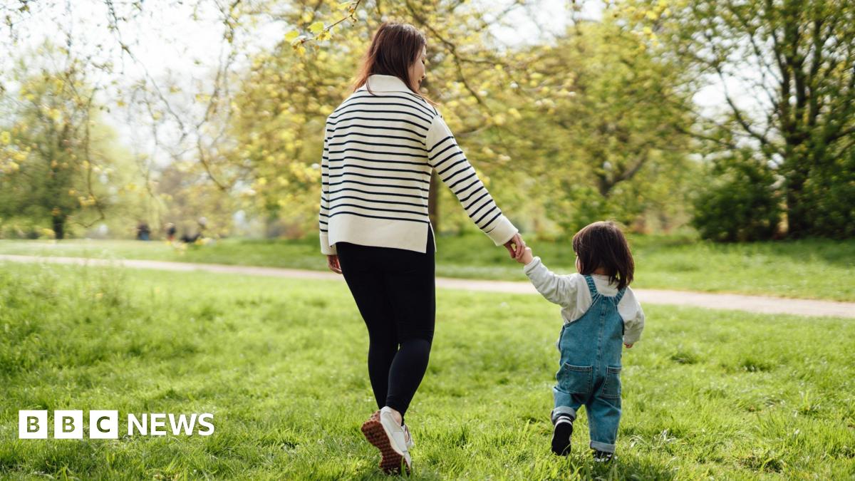 A mum walking and hold her child's hand near trees on a sunny day.