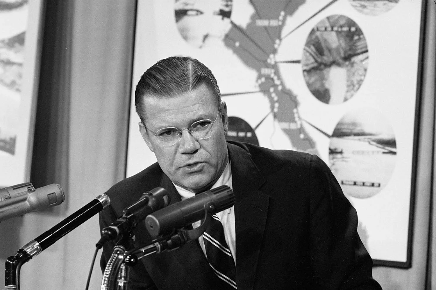 A black-and-white photograph of a man with glasses and slicked-back hair speaking into multiple microphones. Behind him is a large map or diagram featuring circular inset photos.