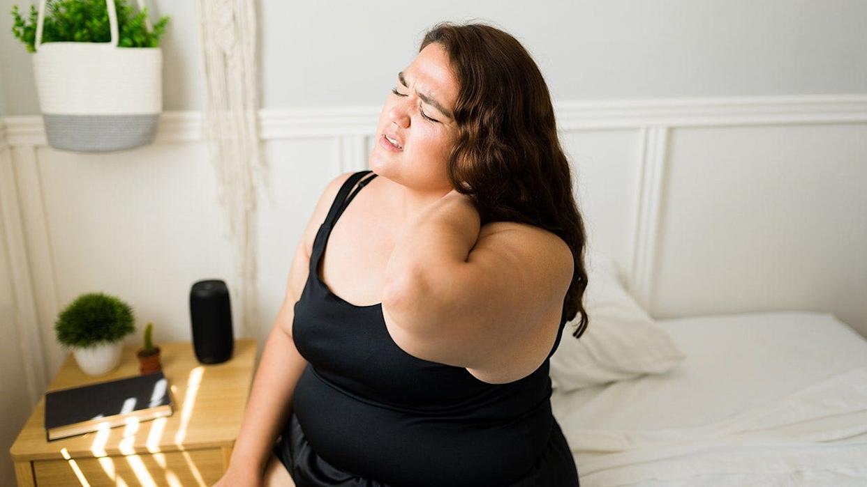 Pained overweight woman rubbing her neck and back in a bedroom.