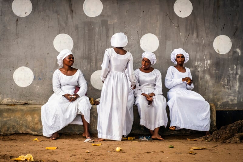 Four women in white dresses and head coverings sit and stand against a gray wall with large white polka dots, engaged in conversation. The ground is sandy with scattered banana peels.