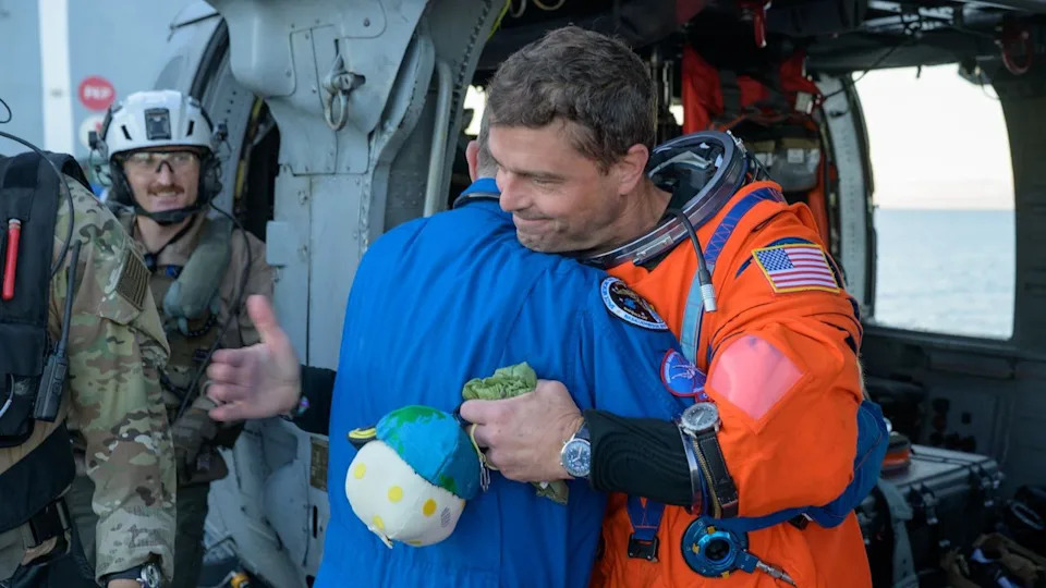  A man in an orange flight suit smiles while carrying a small stuffed moon. 