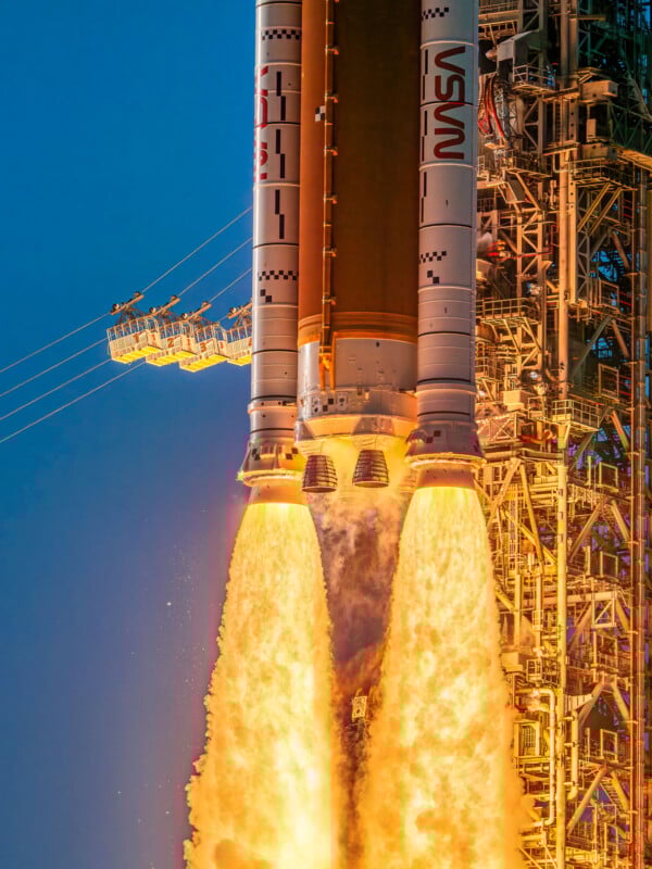 A close-up of a NASA rocket at liftoff, with bright flames and exhaust billowing from its engines, set against a blue sky and surrounded by part of the launch tower.