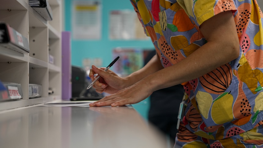 A woman wearing bright, fruit-patterned hospital scrubs leans over with pen and paper