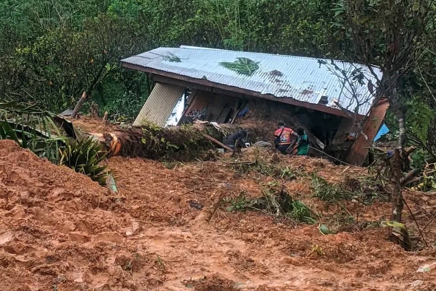 A house sits severely damaged surrounded by mud and debris as two people attempt to look inside it