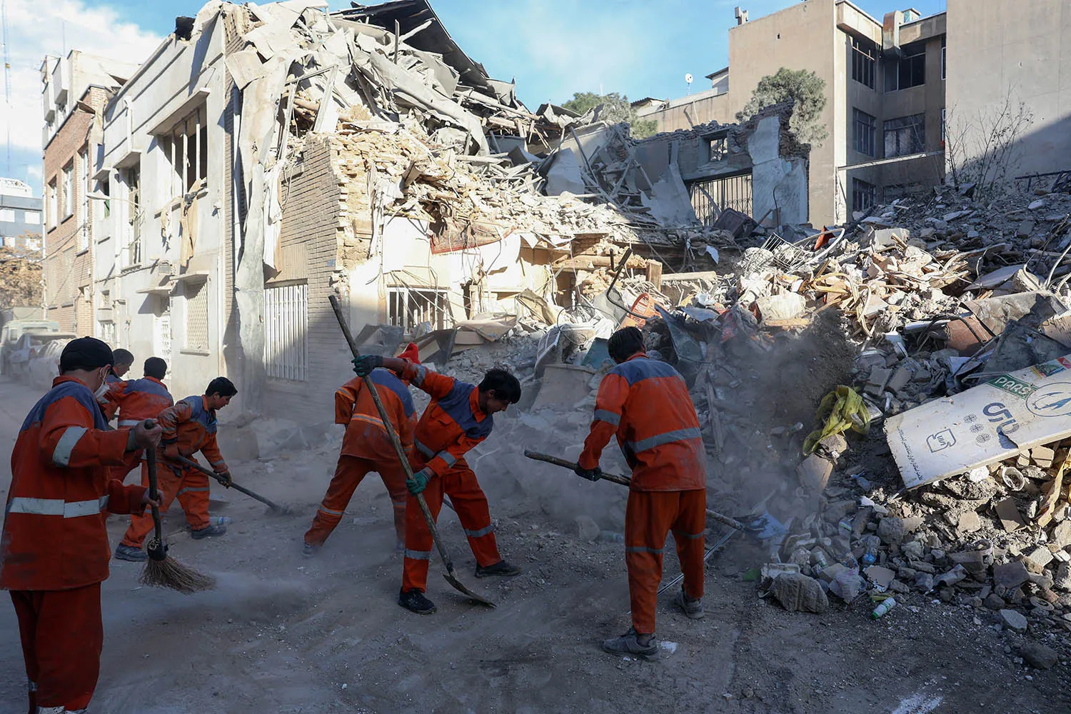 A group of seven workers in bright orange jumpsuits use shovels and brooms to clear dust and debris from a street. In the background, a building has been heavily damaged, with large sections of its structure collapsed into a pile of rubble.