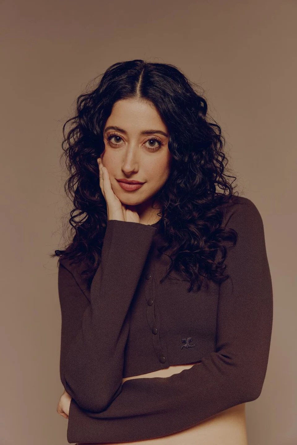 A woman with long curly hair in a long-sleeve brown crop top cardigan holds an arm across her waist and another by her chin.
