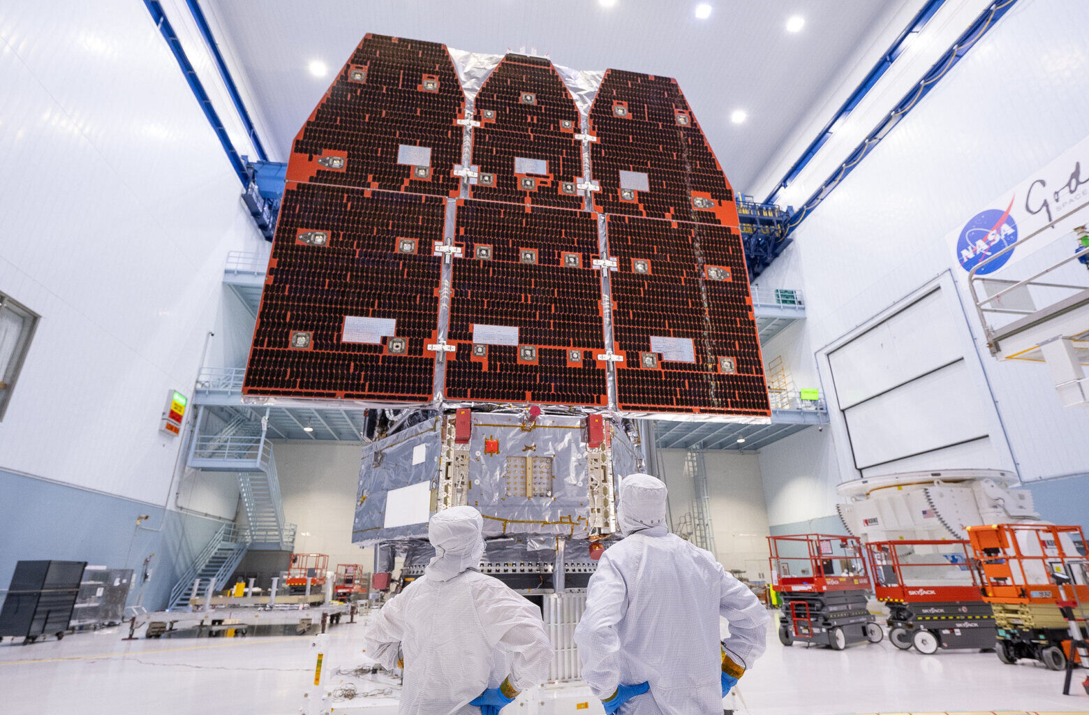 technicians in white body suits and surgical gloves from waist up pictured from behind in foreground stare up at large space telescope in background