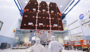 technicians in white body suits and surgical gloves from waist up pictured from behind in foreground stare up at large space telescope in background