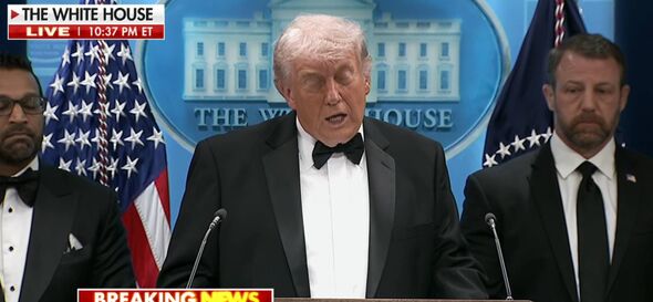 An individual dressed in formal attire, specifically a black tuxedo with a white shirt and a black bow tie, stands at a podium. 