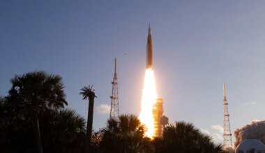 The Artemis II Moon rocket lifts off. There is a bright yellow-orange plume of fire underneath the rocket. The trees in the foreground appear in shadow.