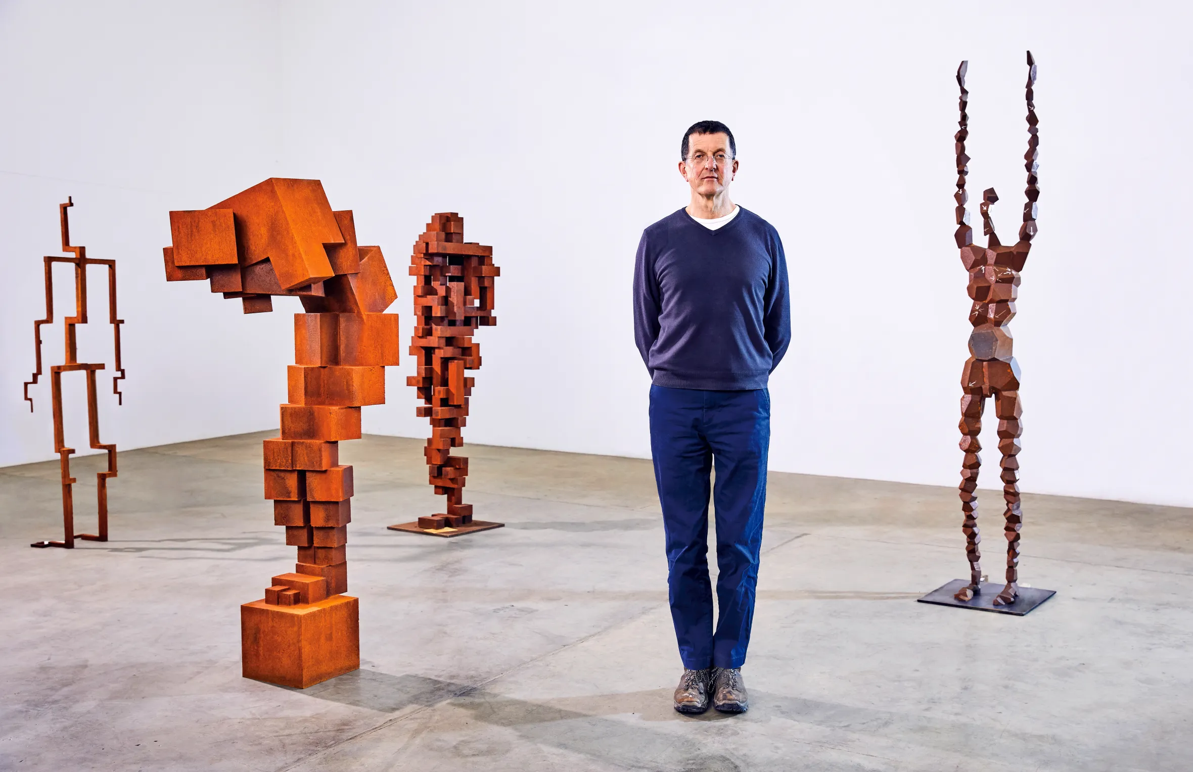 Antony Gormley standing in his studio surrounded by his sculptures, which are abstract human forms made of rusted metal.
