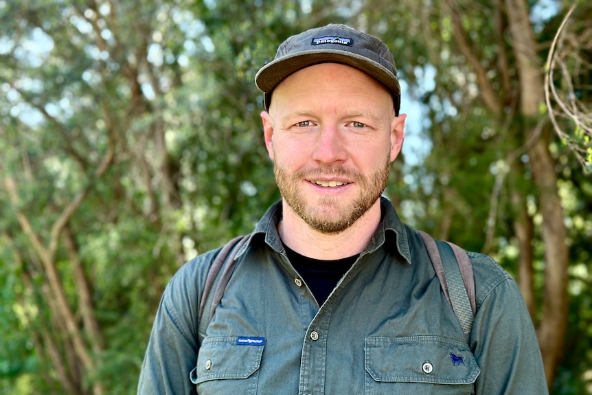 A younger man with a short beard standing in front of trees wearing a cap.