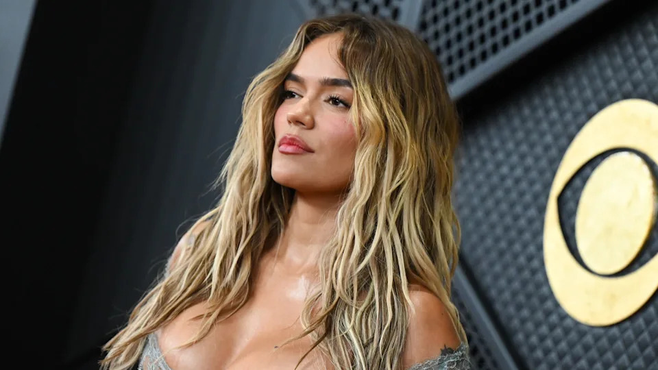 Karol. G with long, wavy hair poses confidently in front of a textured wall.