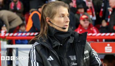 Marie-Louise Eta, wearing a black coat and top with a black microphone and headset in her right ear, on the touchline with fans in red and dark clothing in the background