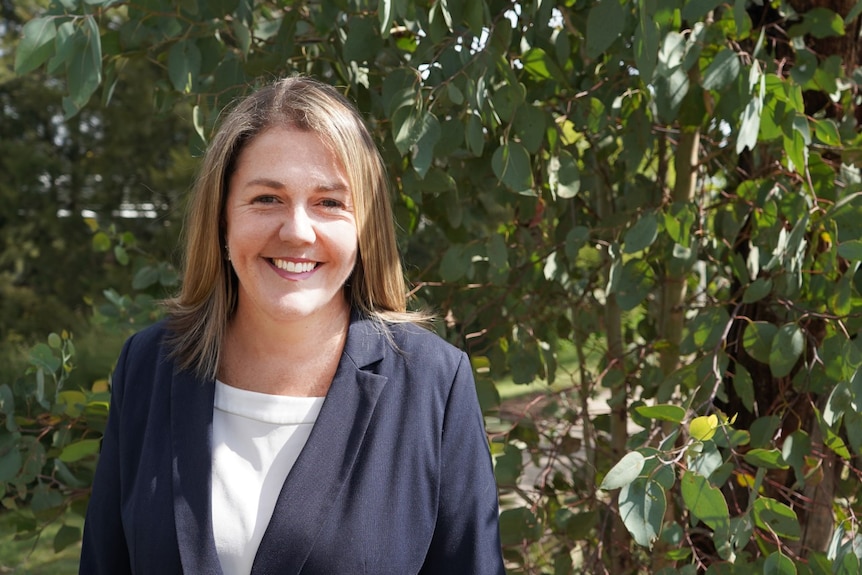 Woman smiling looking at the camera. You can see her upper body and face. She's standing in front of a leafy tree