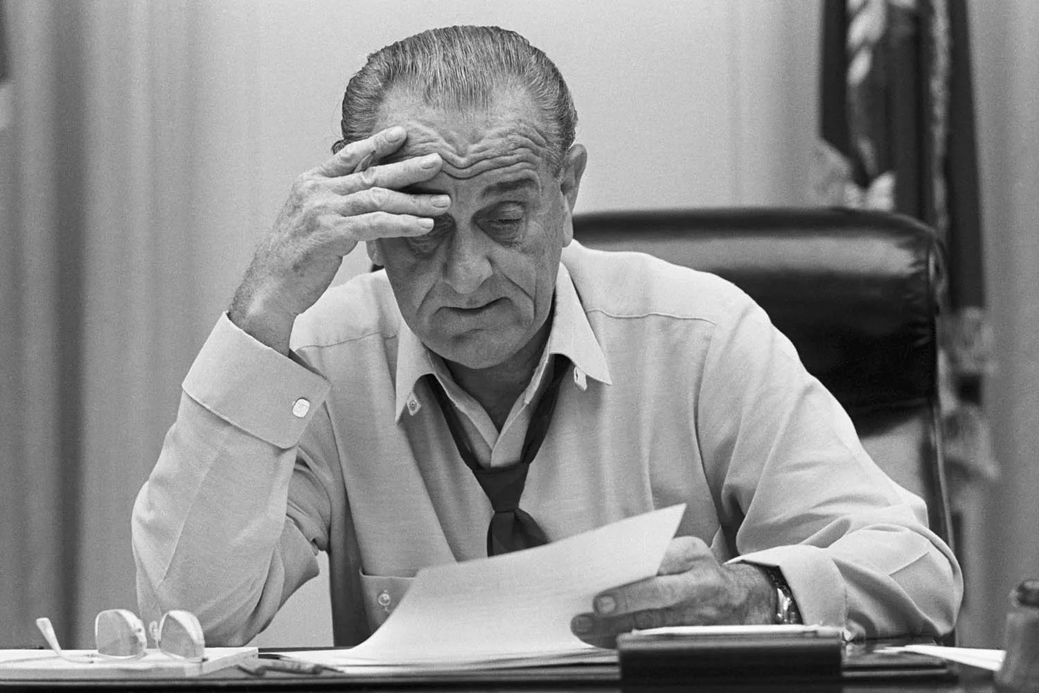 A black-and-white photograph of Lyndon B. Johnson sitting at a desk, looking down at a document with a weary expression. He has one hand pressed against his forehead and his tie is loosened.