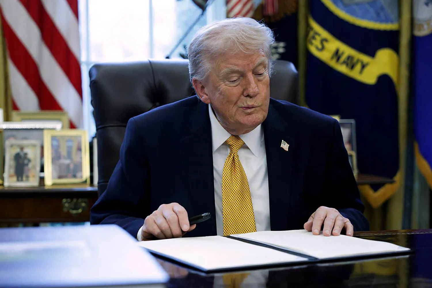 Donald Trump sits at a large wooden desk in the Oval Office, looking down at an open folder. Several framed photos and various flags, including the U.S. Navy flag, are visible in the background.