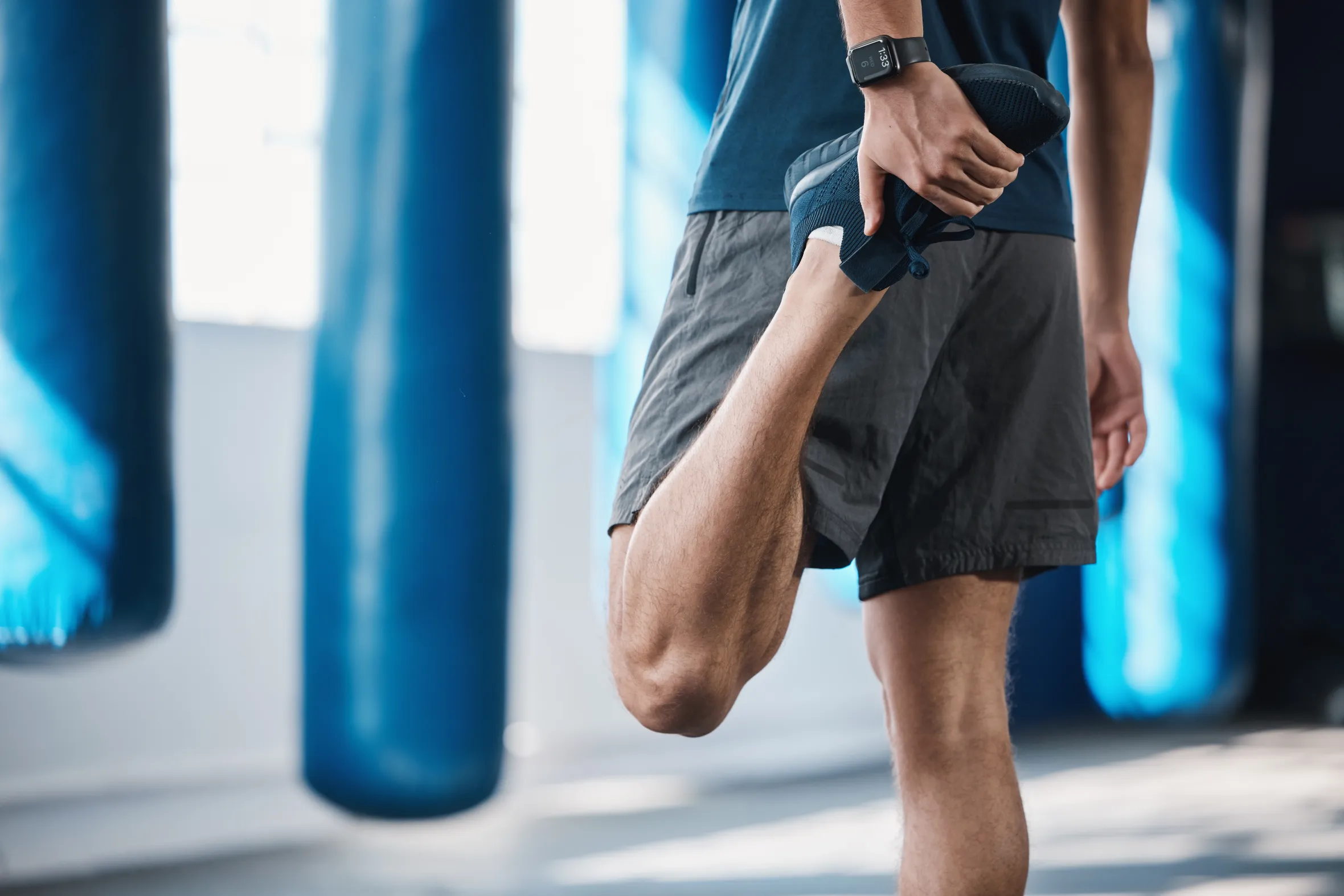 A man stretching his quadriceps in a gym with punching bags in the background.