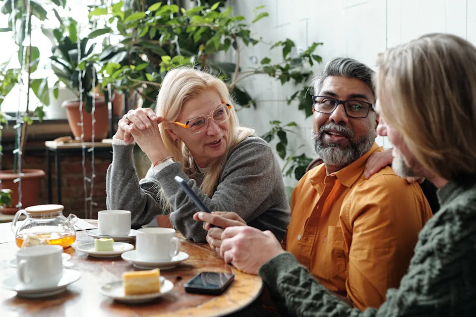 Group of mature friends gathering in cozy cafe, enjoying coffee and pastries while discussing ideas. Hanging out in relaxed, friendly atmosphere with plants and sunlight streaming in