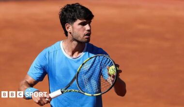 Carlos Alcaraz is wearing a blue t-shirt and holding a tennis ball to his racquet