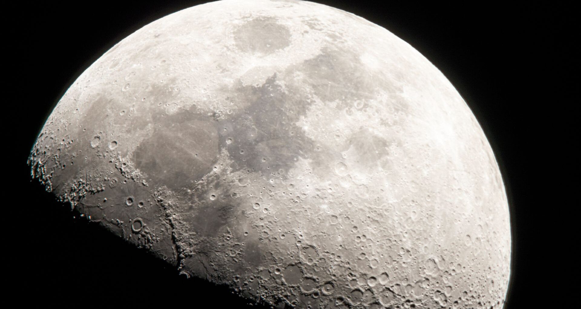 A close up of the moon in the darkness of space, its craters and dark maria visible against the gray surface, while its bottom half is covered in shadow.