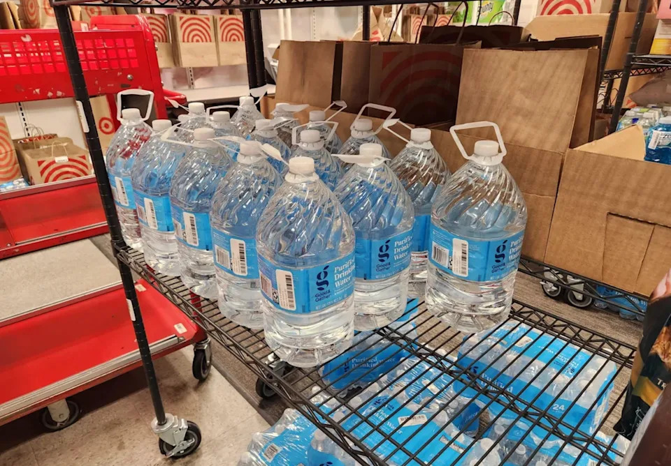 Bottles of water are stocked on metal shelves in a store area, with cardboard boxes and shopping carts visible in the background