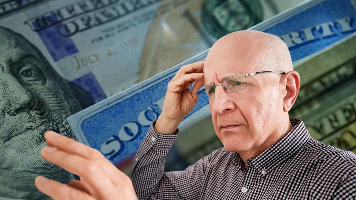 A close-up of a bald, older man with glasses, wearing a checkered shirt, holding his hand to his forehead with a thoughtful or worried expression. The background is blurred, showing a portion of a U.S. $100 bill with Benjamin Franklin's face and blue strips resembling a Social Security card with text like "SOCIAL" and "RITY" visible.