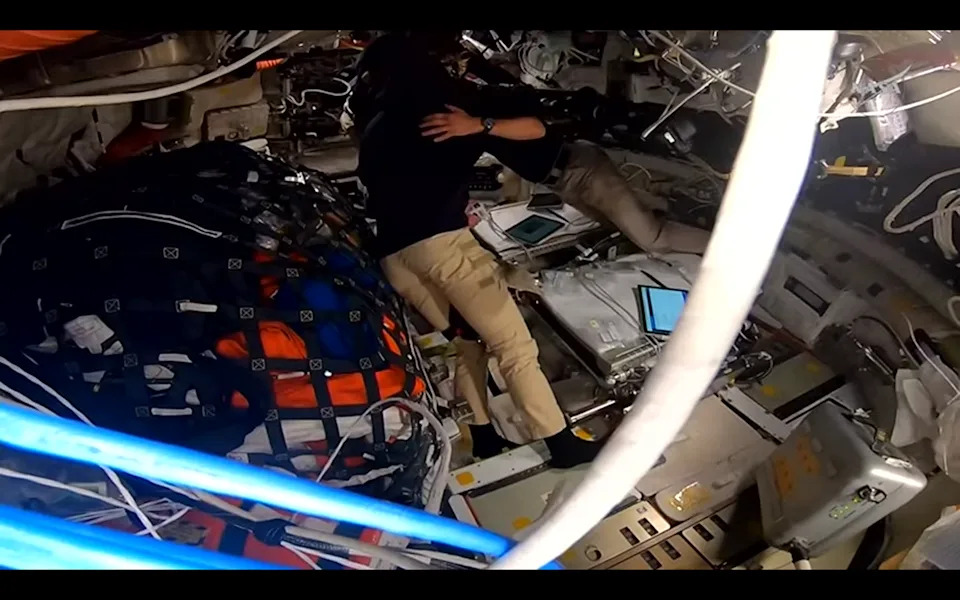 four people hug inside a cramped spacecraft full of wires and cargo under netting