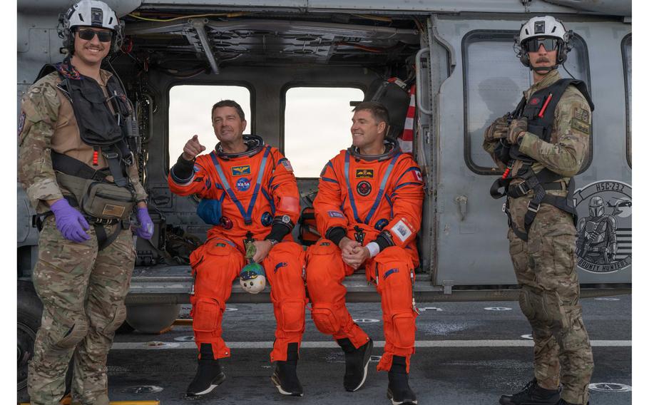 Two astronauts in orange space suits sitting in a helicopter, sandwiched between two service members.