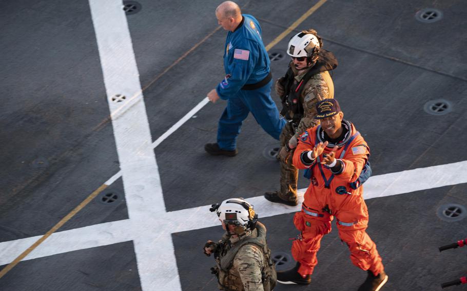 An astronaut in a baseball cap walking on the deck of a ship next to sailors.