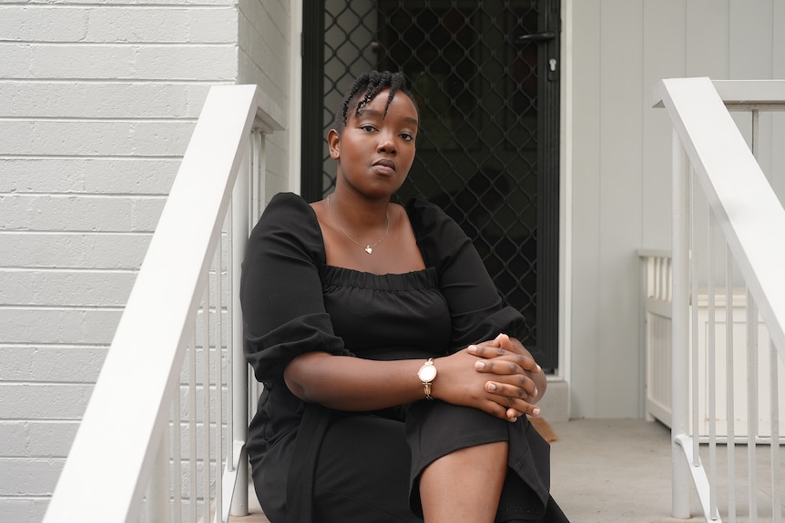 A Kenyan-born woman in her 20s wearing a black dress sits on the steps outside her white brick house.