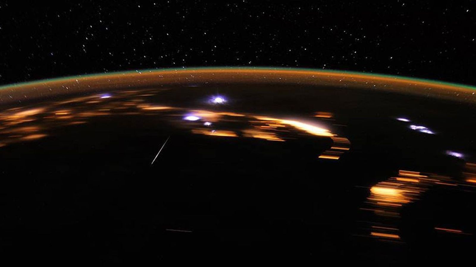A view of the Earth from space, its dark surface showing glowing city lights with sparse purple glowing areas where meteors hit the atmosphere.
