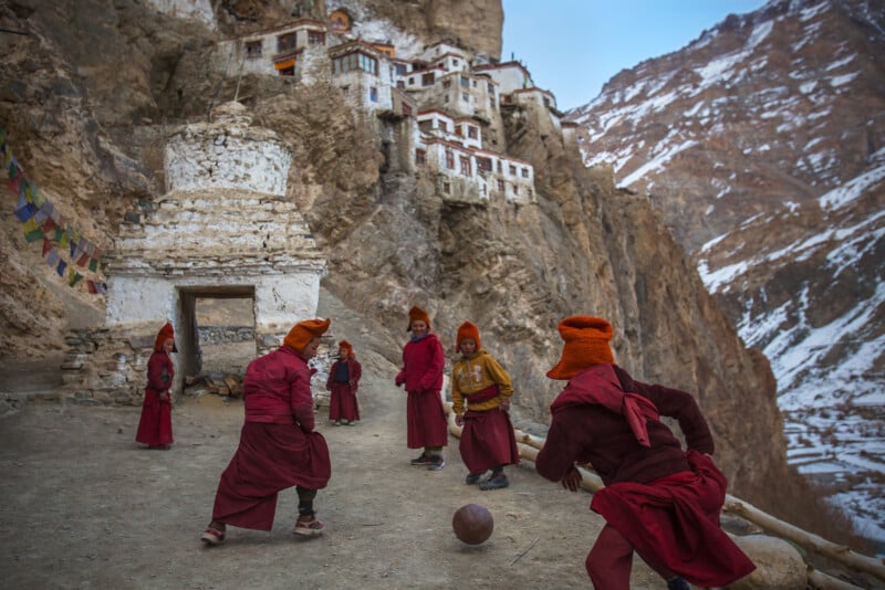 A group of young monks in red robes and orange hats play with a ball outside a monastery built into a rocky, snowy mountainside. Colorful prayer flags hang nearby.