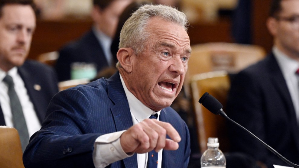 Robert F. Kennedy Jr. speaks into a microphone during a hearing with members of the US House. His left hand is loosely cupping a water bottle and his right arm is lifted to emphasize his speech. He is wearing a blue suit.