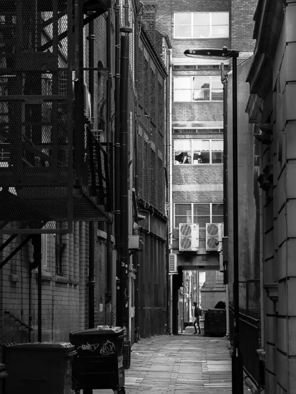 A narrow urban alleyway with tall buildings on both sides, vents and fire escapes, rubbish bins, and a single person walking in the distance; the photo is in black and white.
