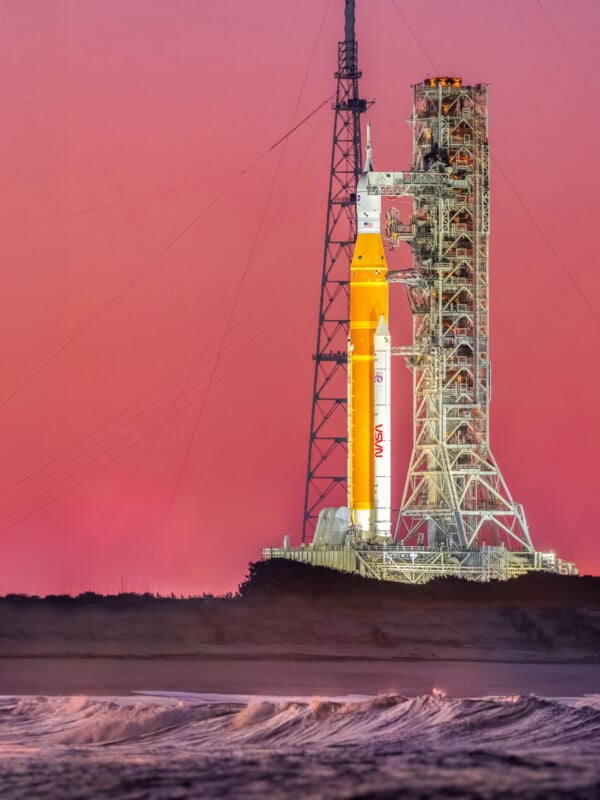 A NASA rocket stands vertically on a launch pad beside a tall metal structure at sunrise or sunset, with waves from the ocean in the foreground and a pink sky in the background.