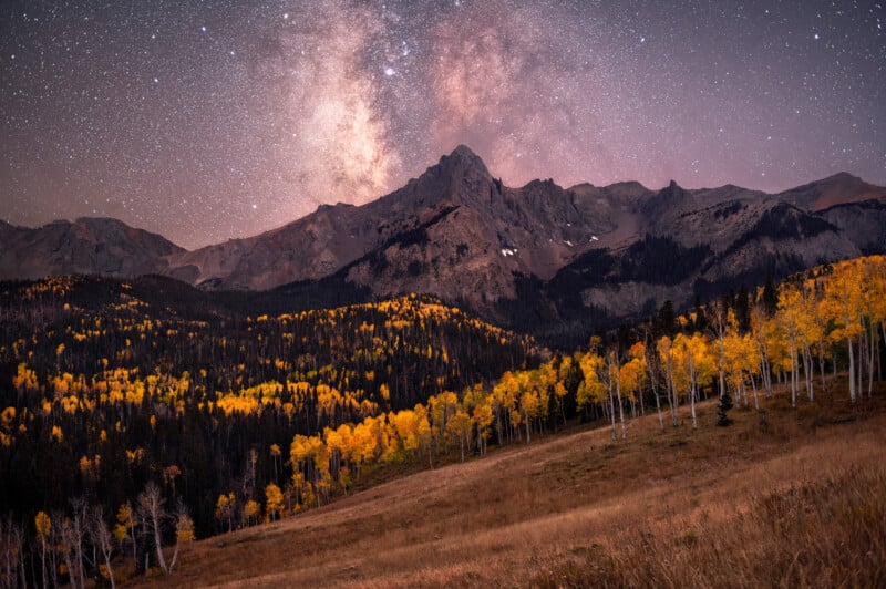 A mountain range towers above a colorful autumn forest with yellow trees in the foreground, under a star-filled night sky featuring the bright core of the Milky Way.
