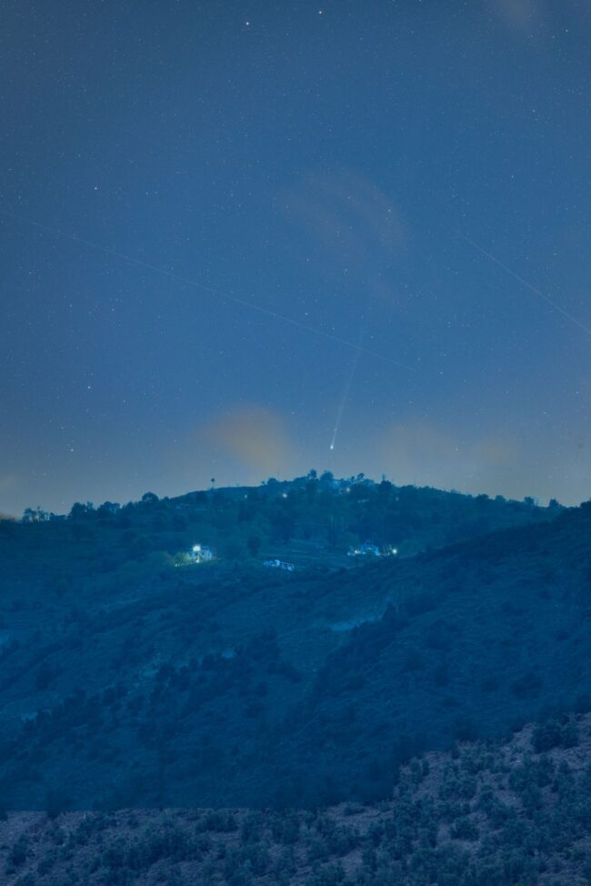 A blue-tinted night scene with some lights of civilization on the mountainside and a comet above.