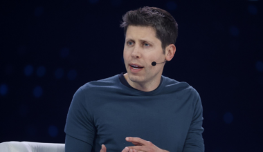 A man wearing a dark blue long-sleeve shirt sits in a white chair, speaking with hand gestures and wearing a headset microphone.