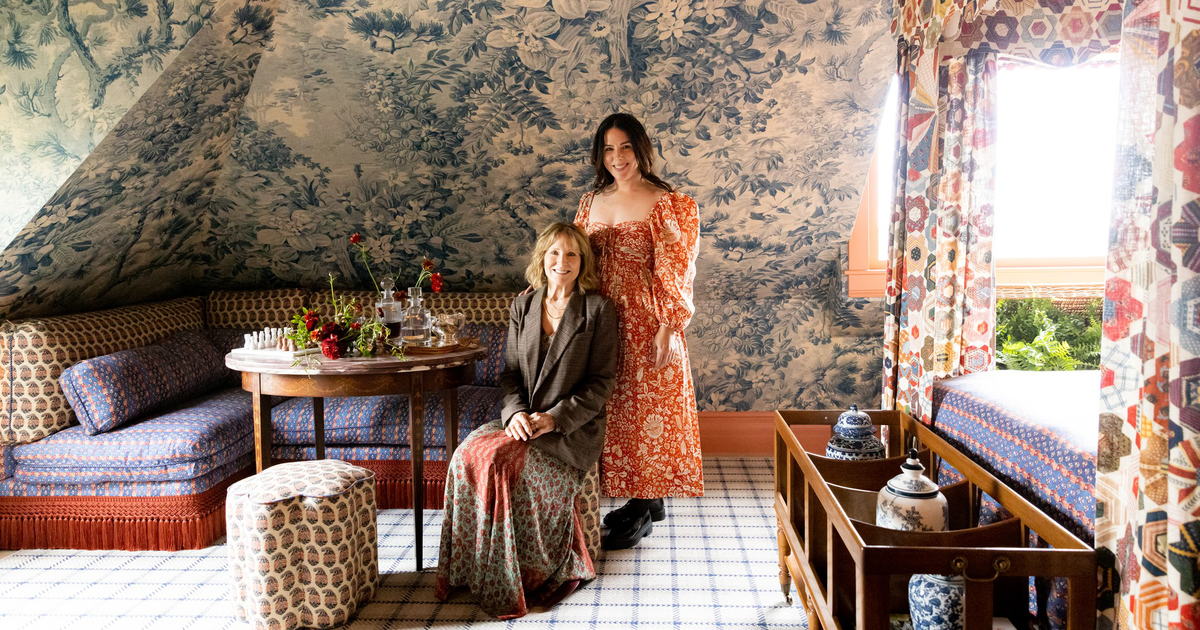 Two women pose in a cozy, patterned room with floral wallpaper, a wooden table set with flowers, and colorful curtains framing a bright window.