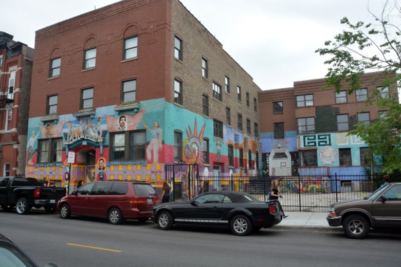 A street view of the Casa Aztlan mural, which stretches around an L-shaped brick building. Cars parked on the street partially obscure the view. The mural is painted in swatches of turquoise, purple, pink, and yellow. and depict people and imagery related to Mexican history.