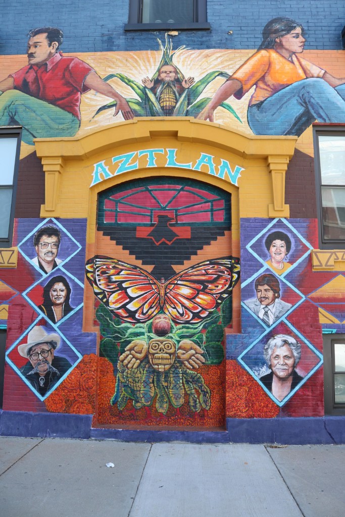 A colorful mural on a brick building in Pilsen. The word Aztlan is painted in blue above a bricked entryway. The door features a butterfly below an inverted pyramid and above a skull and hands. Surrounding the doorway are images of people.