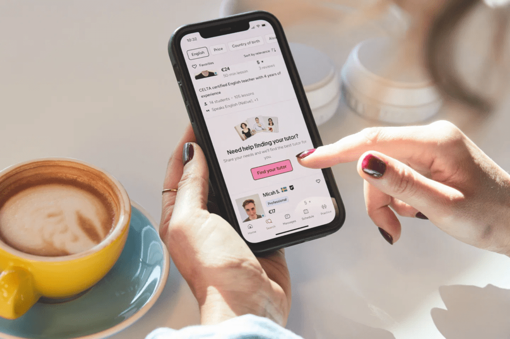 A hand holding a phone displaying a tutor search app with a "Find your tutor" button and a coffee cup in the foreground.