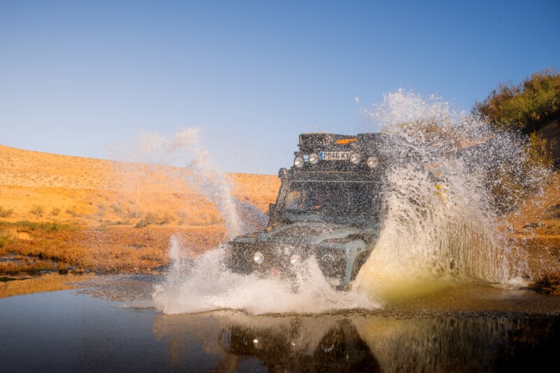A rugged off-road vehicle drives through a shallow water stream in a desert landscape, creating a large splash. The sunlit sand dunes and clear blue sky are visible in the background.