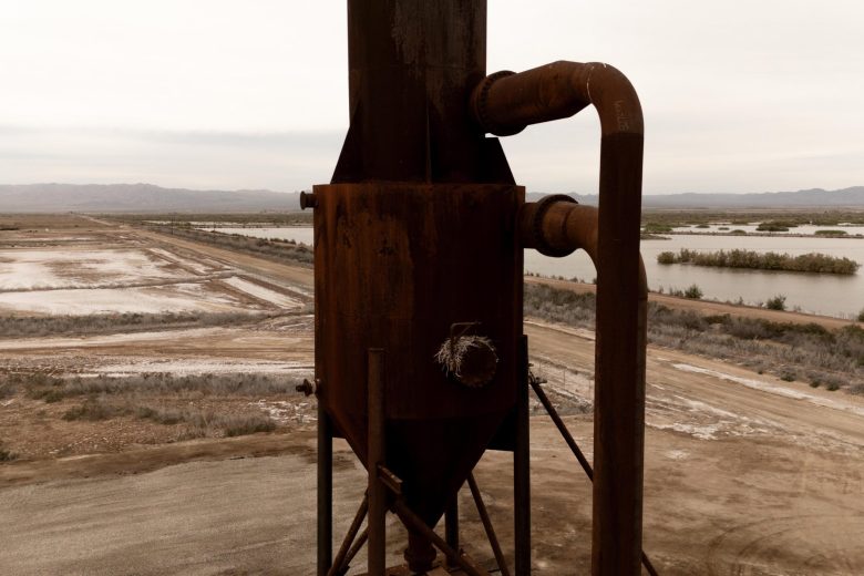 A rusted industrial structure with pipes at a geothermal site near the Salton Sea, with arid landscape and water channels in the background.