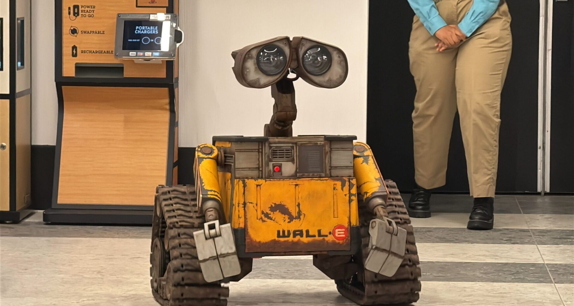 A WALL-E robot replica stands on a tiled floor indoors near a person in khaki pants and blue shirt.