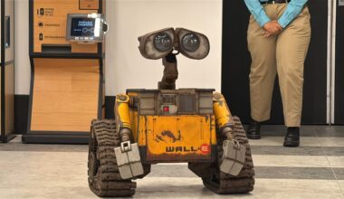 A WALL-E robot replica stands on a tiled floor indoors near a person in khaki pants and blue shirt.