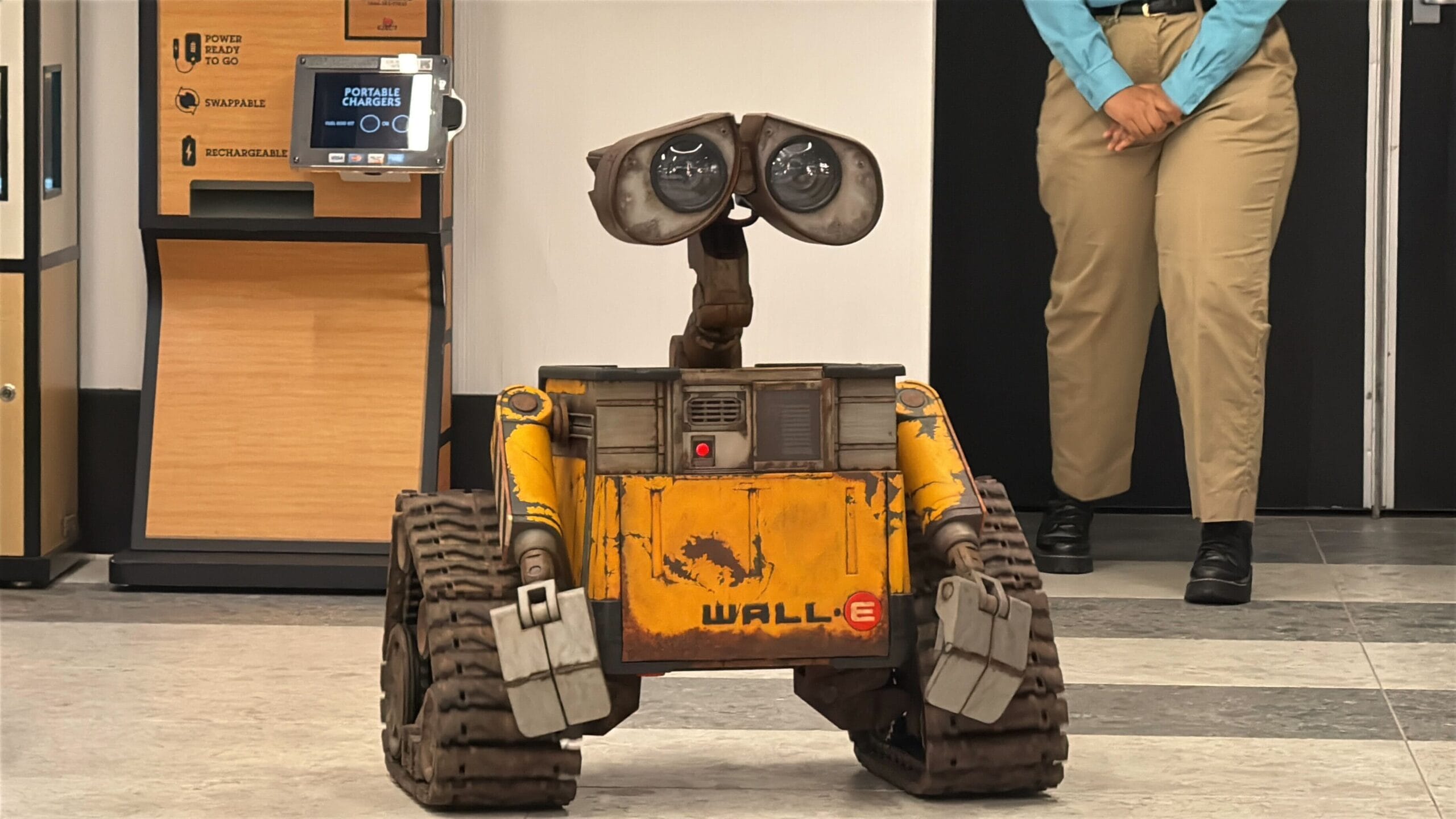 A WALL-E robot replica stands on a tiled floor indoors near a person in khaki pants and blue shirt.