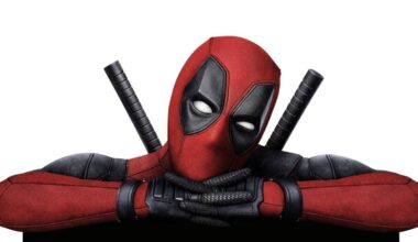 A person in a red and black Deadpool costume with a mask and two swords on their back poses with crossed arms.